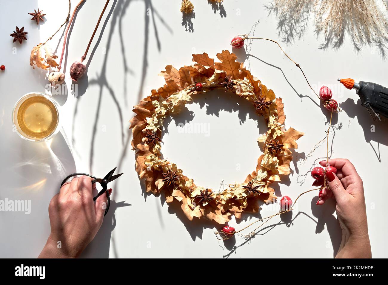 Hands making dried floral wreath from dry Autumn leaves and Fall berries. Hands with manicured nails fix decorations with glue gun. Stock Photo