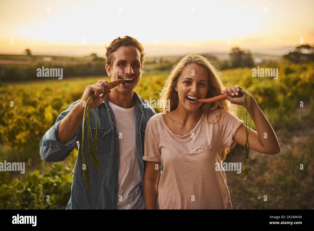 Just testing the produce. Shot of a young couple each holding up a carrot and taking a bite with their crops in the background. Stock Photo