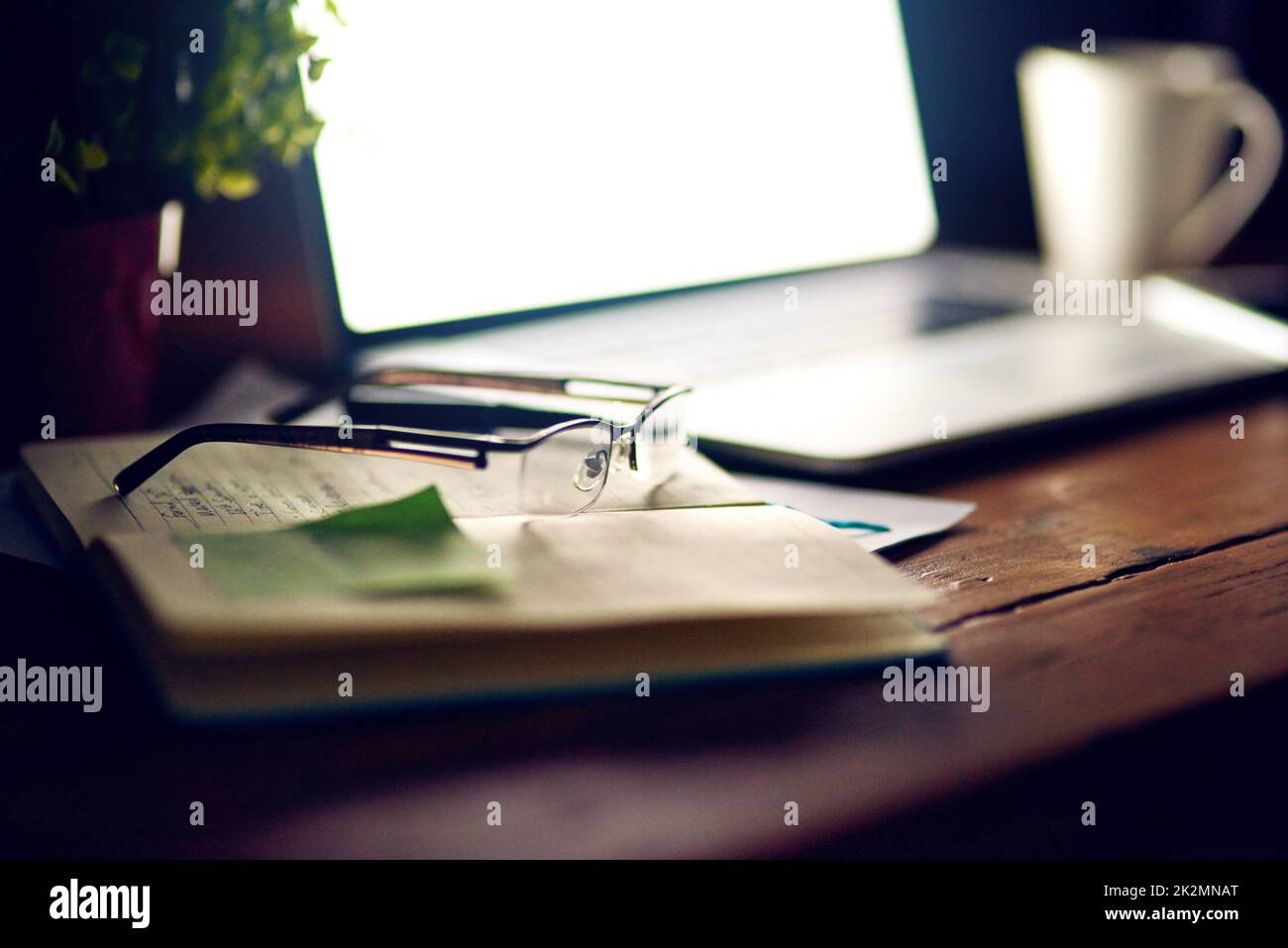Coming back to it in the morning. Shot of reading glasses on top of a notepad and a laptop on a desk. Stock Photo