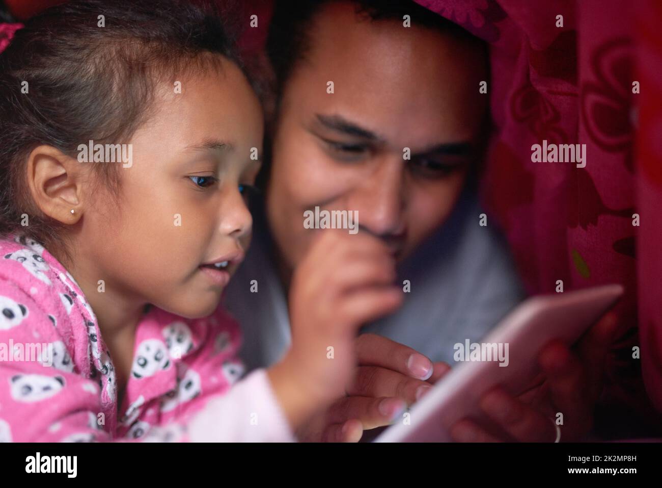 Story time is her favorite part of going to bed. Shot of a father reading a story to his little girl from a digital tablet while lying under together under a blanket. Stock Photo