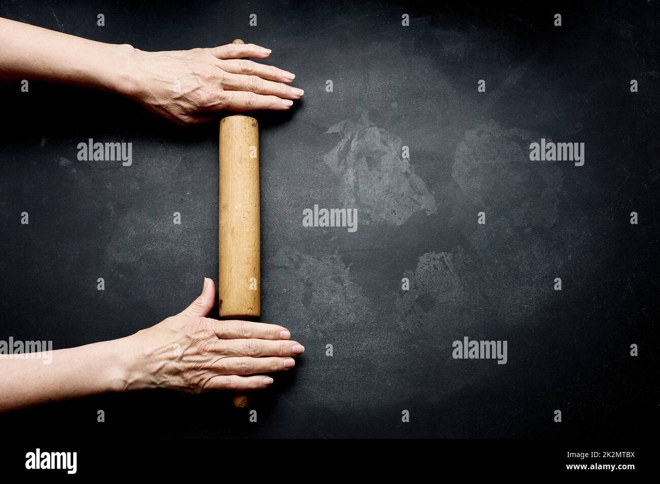 two female hands hold a wooden rolling pin for rolling dough on a black table, top view. Stock Photo