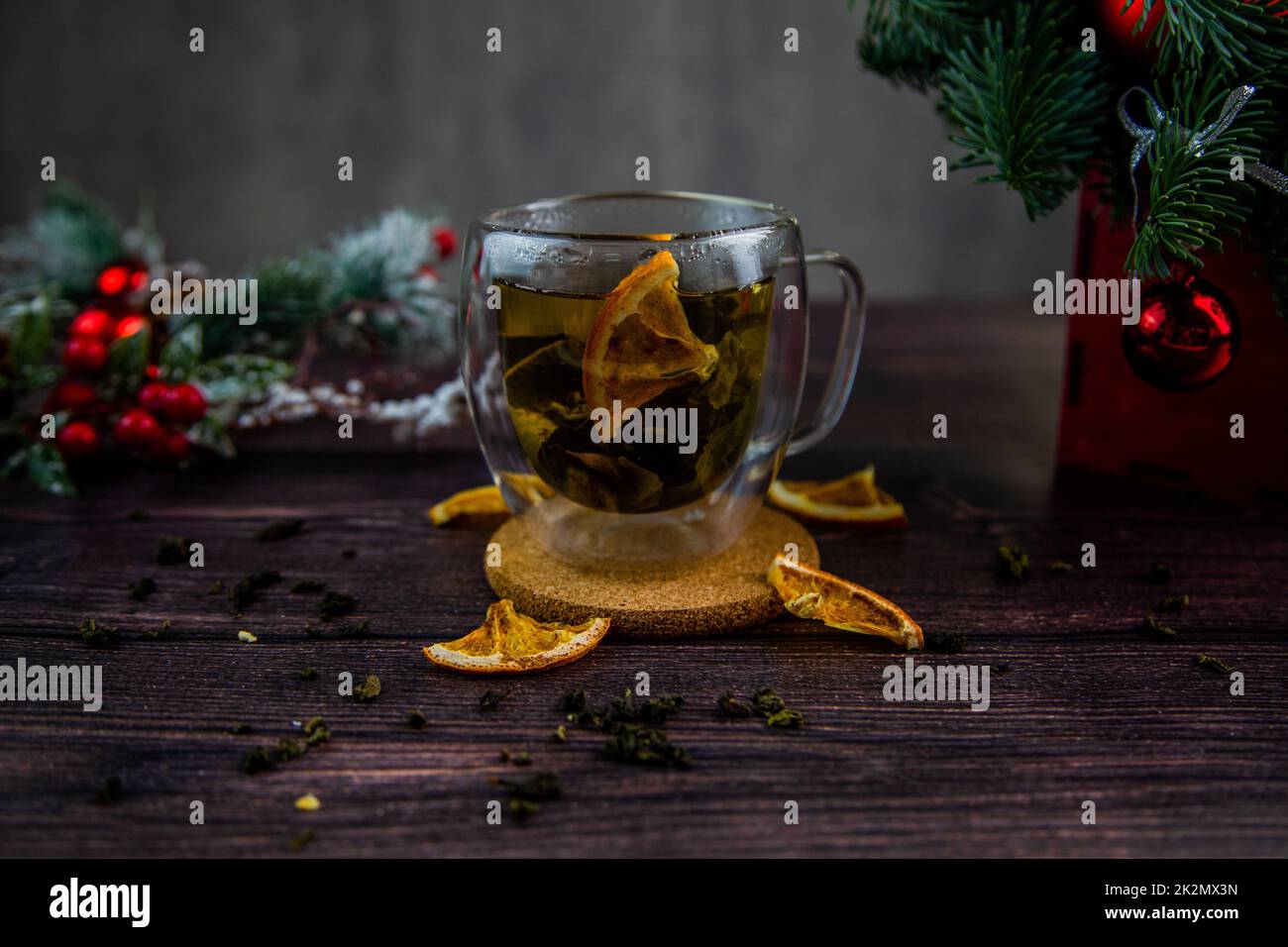 A transparent cup of tea stands on a dark table, on a wooden stand, dried orange slices float in the cup, citrus slices lie next to it, tea is scattered, a photo in a dark key Stock Photo