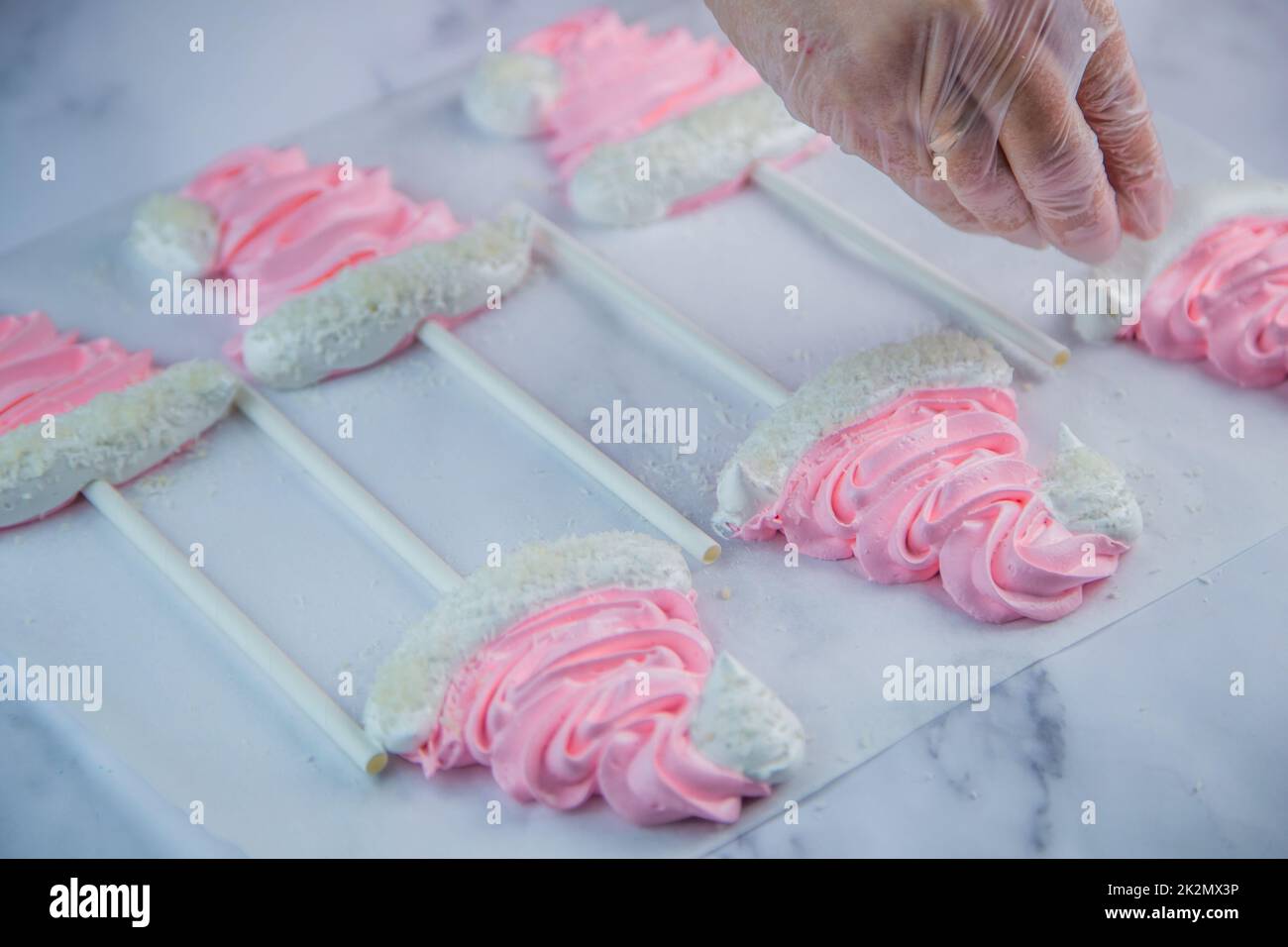 A pastry chef's hand in a transparent glove sprinkle coconut shavings on a pink meringue cap on a stick that lies on white parchment Stock Photo