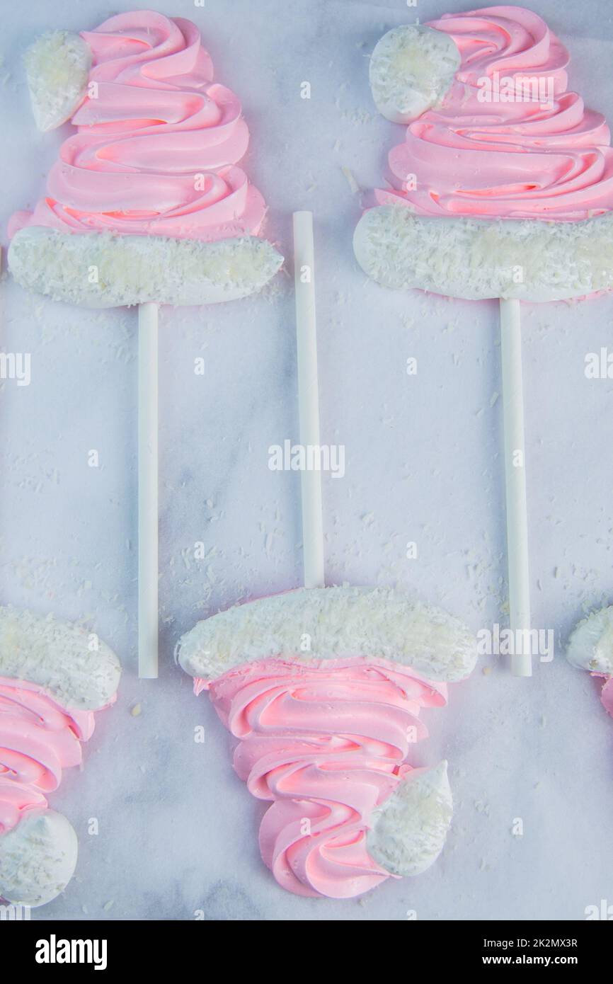 Meringue in the form of pink Christmas hats with a pompom and a white lapel on a stick lie on white parchment in two rows Stock Photo