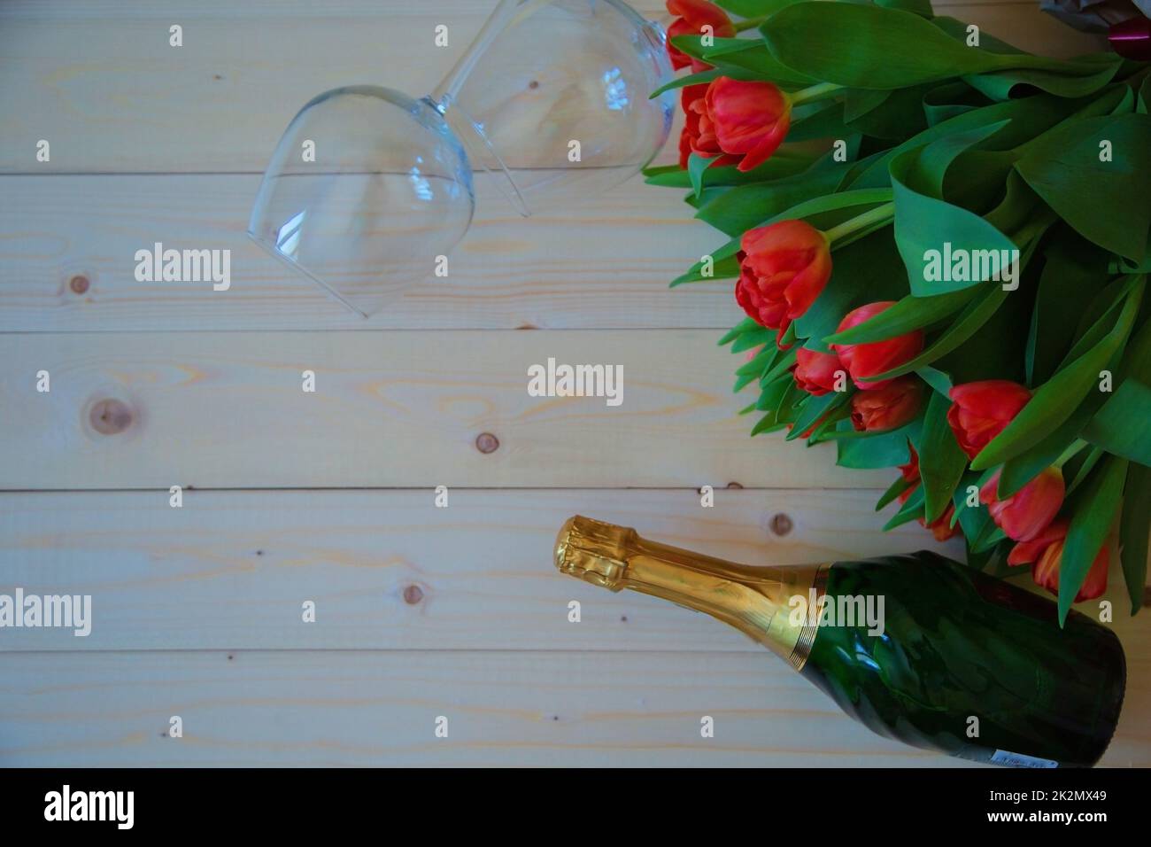 Pink tulips with green leaves lie in the upper corner on a wooden background, and on the sides of the bouquet are wine glasses and sparkling wine Stock Photo