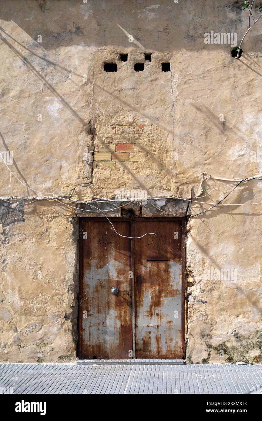 An old metal door with peeling paint on an old building with a white wall and electricity cables pinned to the wall. Stock Photo