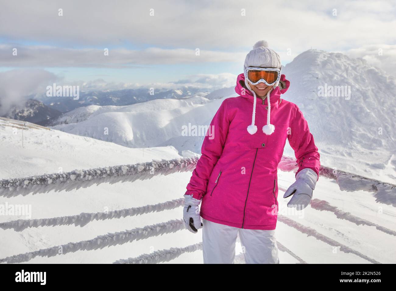 Young woman in pink jacket, wearing ski goggles, leaning on snow covered fence, smiling, white mountains in background. Stock Photo