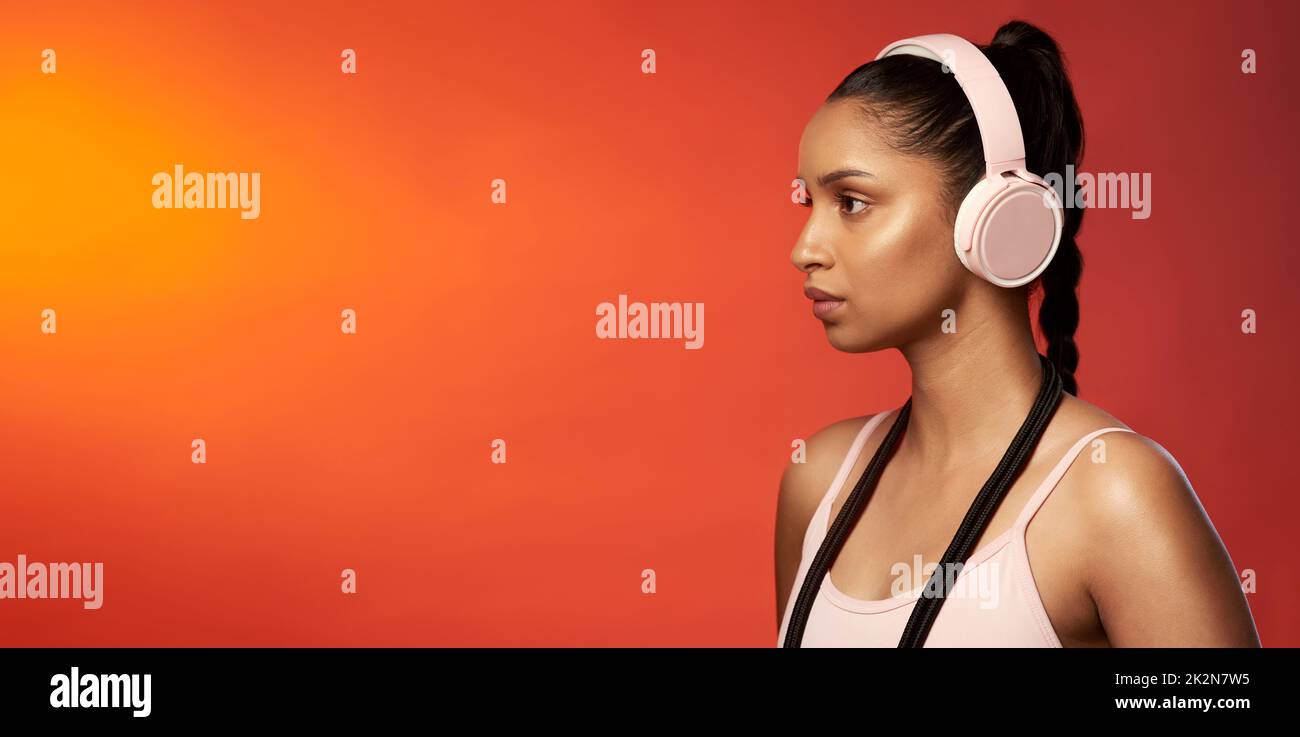 Playing tracks that keep her focused. Studio shot of a sporty young woman wearing headphones and posing with a skipping rope around her neck against a red background. Stock Photo