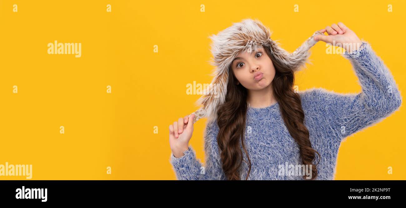 smiling child in sweater and earflap hat on yellow background, wintertime. Banner of child girl in winter hat, studio poster header with copy space. Stock Photo
