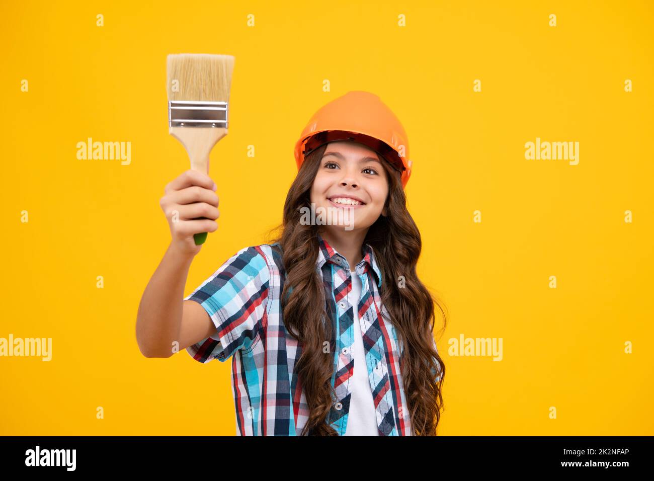 Painter in workshop. Renovation and repair. Teen girl in helmet with painting brush. Child in hard hat. Kid builder painter on construction site. Stock Photo