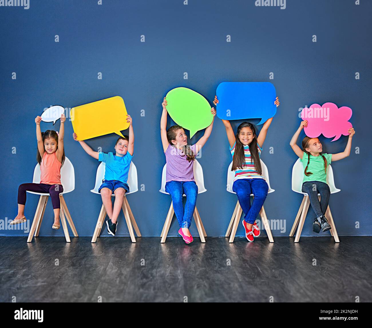 Fill the world with your voice. Studio shot of a diverse group of kids sitting on chairs and holding up speech bubbles against a blue background. Stock Photo
