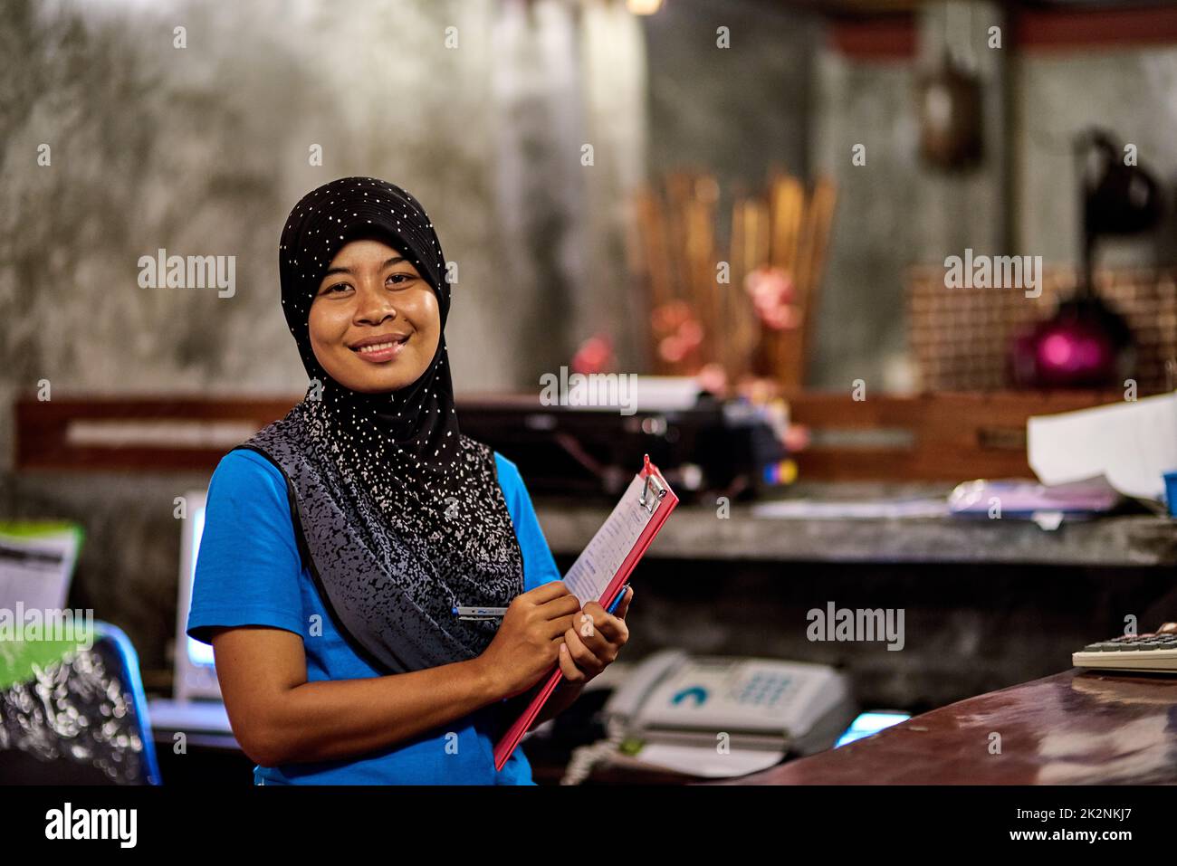 Youre always welcome here. Portrait of a smiling muslim thai woman standing behind a hotel reception desk holding a clipboard. Stock Photo