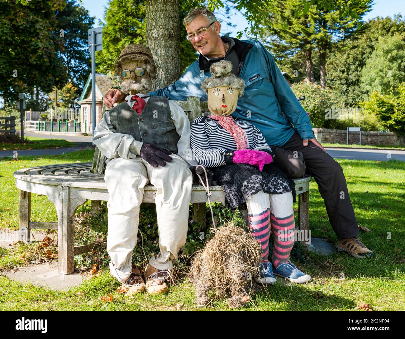 Dirleton, East Lothian, Scotland, UK, 23rd September 2022. Scarecrow Trail: the second trail this year has 18 scarecrows around the village. Pictured: a man sitting with a couple of fabulous scarecrows on the village green. Credit: Sally Anderson/Alamy Live News Stock Photo