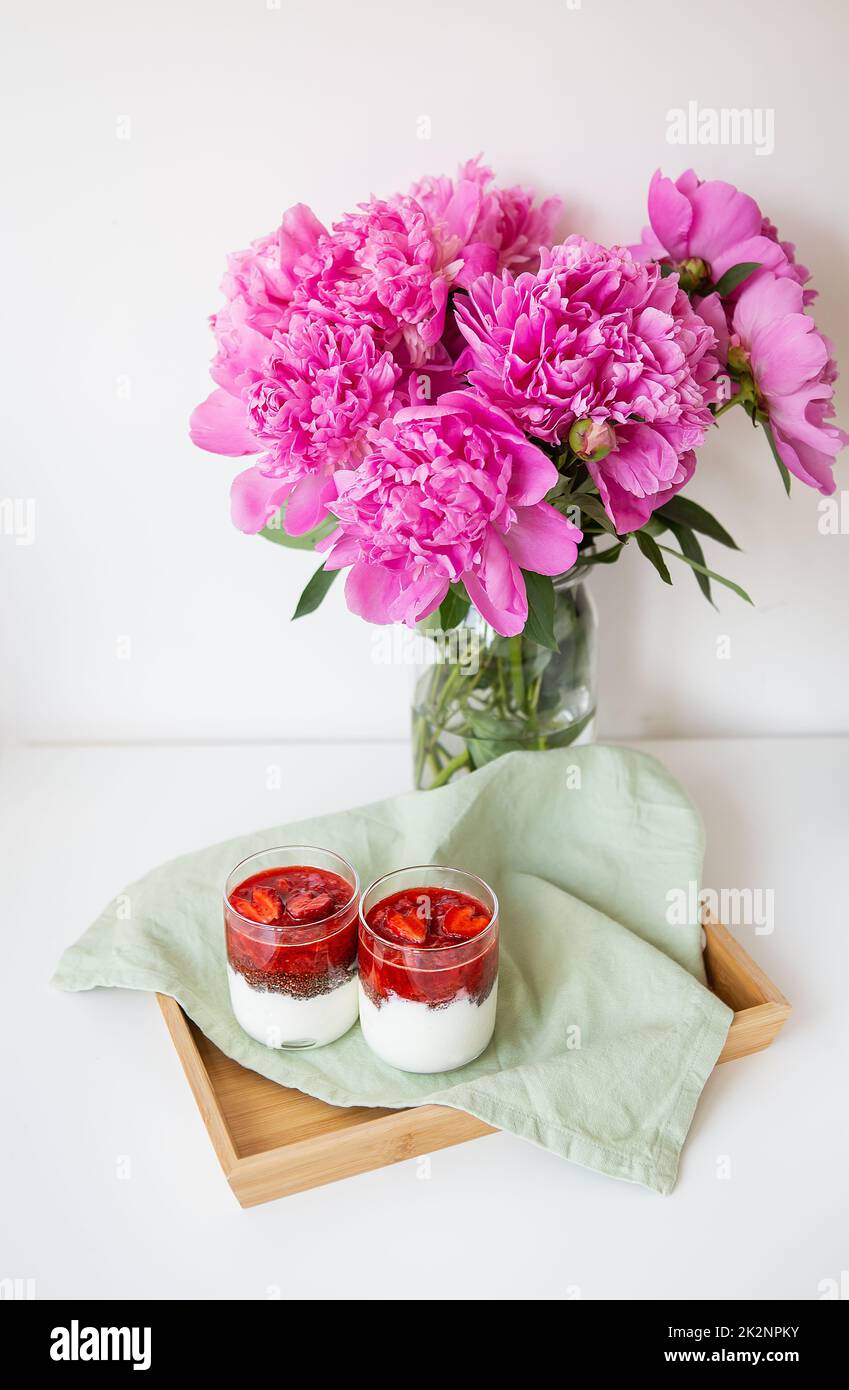 A beautiful bouquet of pink peonies in a vase on a white table stands along with a heart-shaped strawberry dessert. St. Valentine's Day, 8 March. Stock Photo
