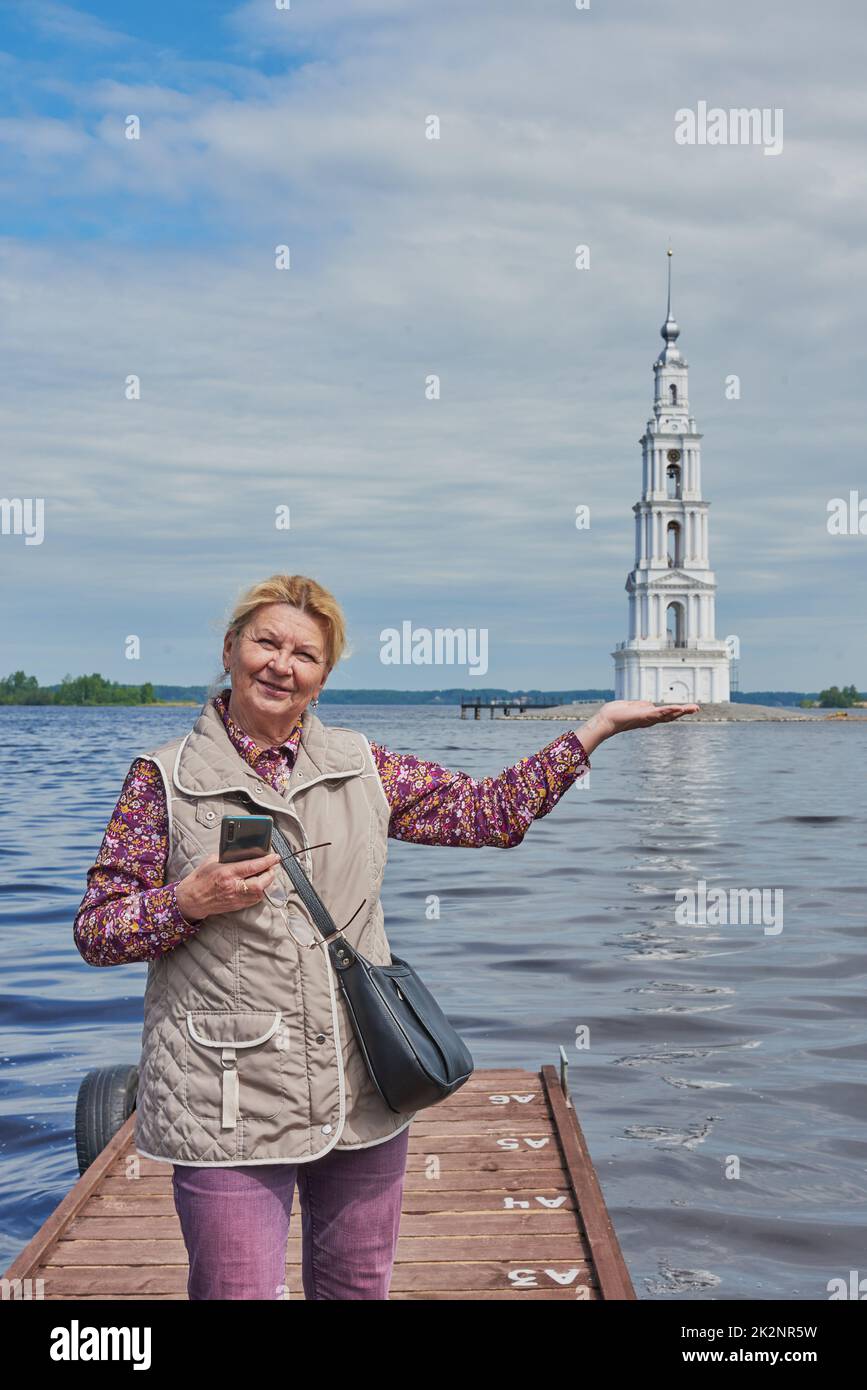 An elderly tourist poses against the background of the bell tower and pretends to hold the tower in the palm of her hand.  Stock Photo