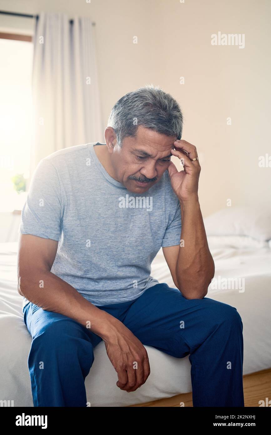 Okay I have to think long and hard about this one. Shot of a stressed out mature man holding his head in discomfort while being seated on his bed at home. Stock Photo