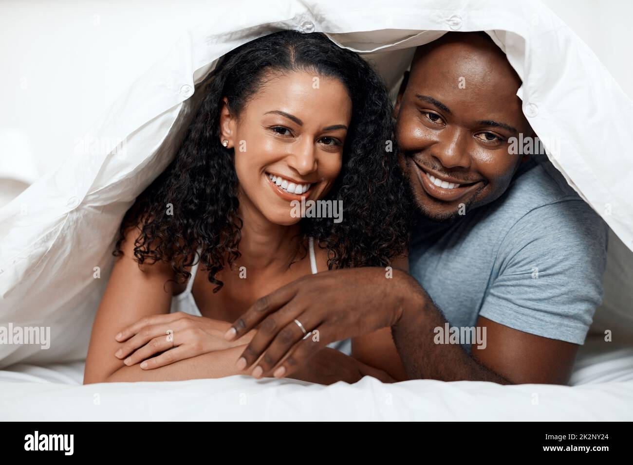 Always in a good mood together. Portrait of a cheerful young couple lying in bed together under a blanket while looking at the camera at home in the morning. Stock Photo
