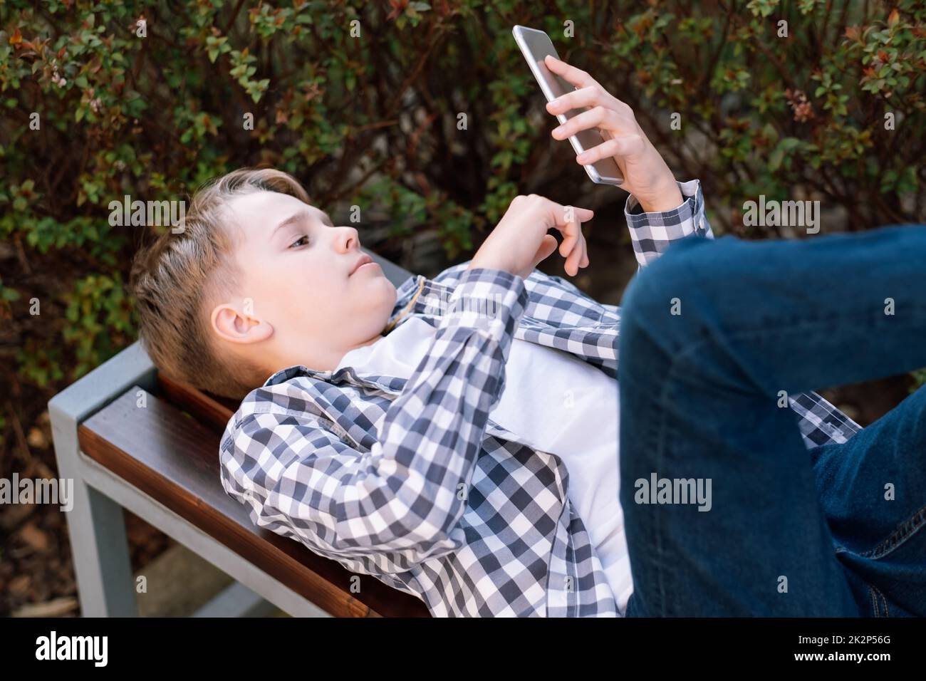 Attractive and thoughtful teenager boy resting lying on bench, holding and using smartphone for networking. Stock Photo