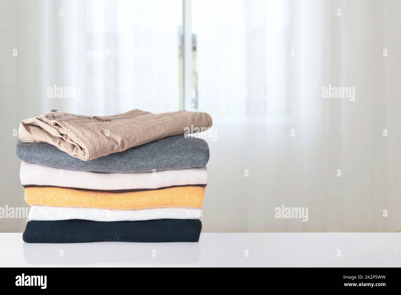 Stack of spring clothes. Pile of colorful knitted cozy warm cashmere sweaters or pullover on table against abstract bright curtain background. Space for display product montage. Beautiful backdrop. Stock Photo