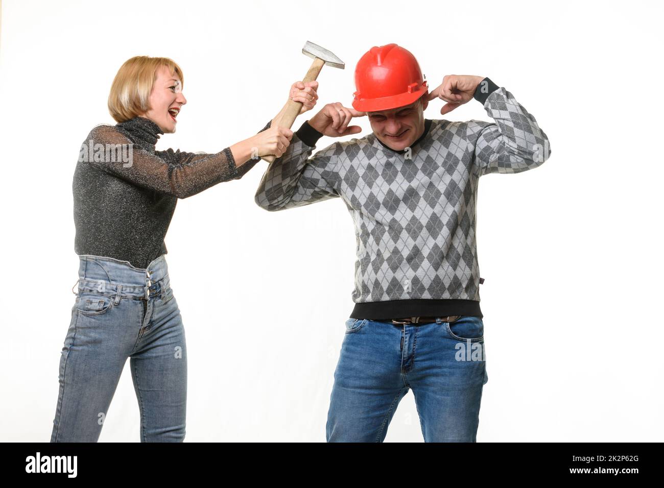 The wife screams and hits her husband on the head with a hammer, the husband is wearing a helmet and he covered his ears with his fingers Stock Photo