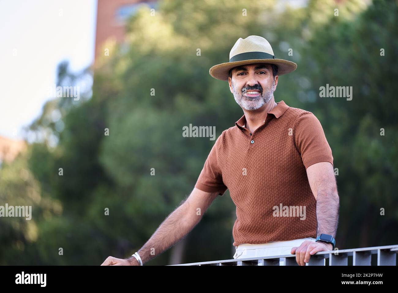 Middle-aged man in hat looking at camera and smiling while leaning on a railing outdoors. Stock Photo
