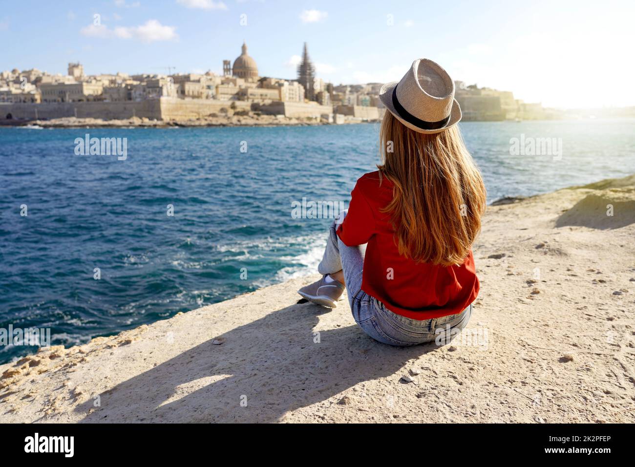 Girl sits down on the edge of the sea enjoying amazing landscape of Valletta, Malta. Travel concept with independent people doing outdoor leisure activity and wanderlust life. Stock Photo
