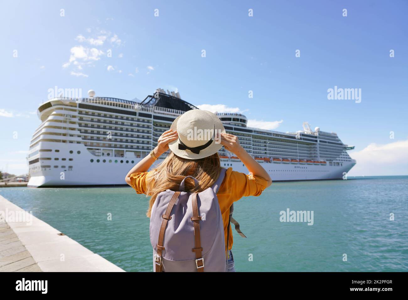 Young female backpacker holding straw hat standing in front of big cruise liner Stock Photo