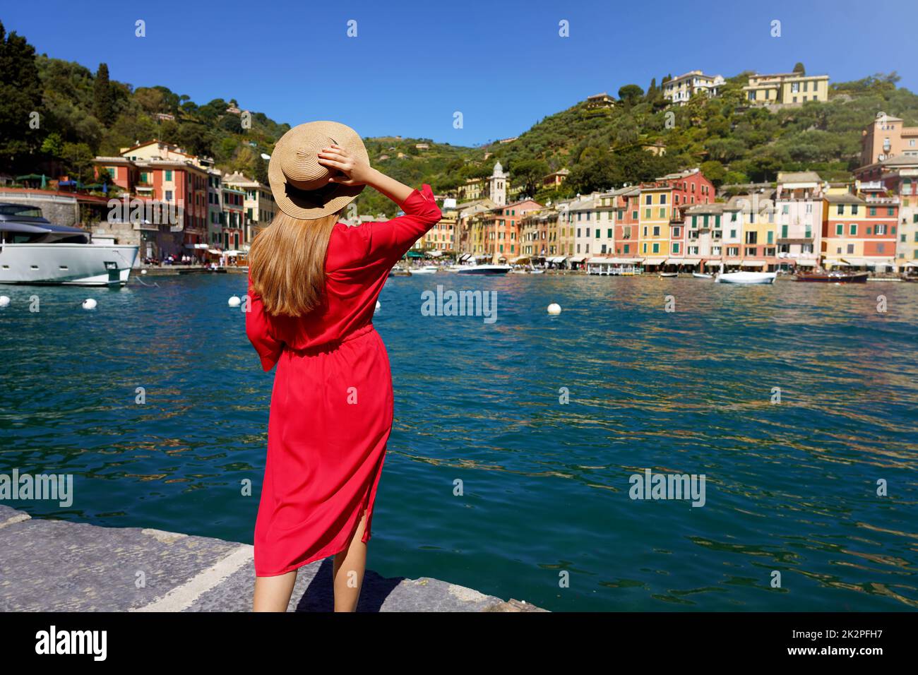 Holidays in Italy. Rear view of beautiful young woman in red dress looks towards Portofino traditional colorful village with yachts moored in its picturesque cozy harbor, Portofino, Liguria, Italy. Stock Photo