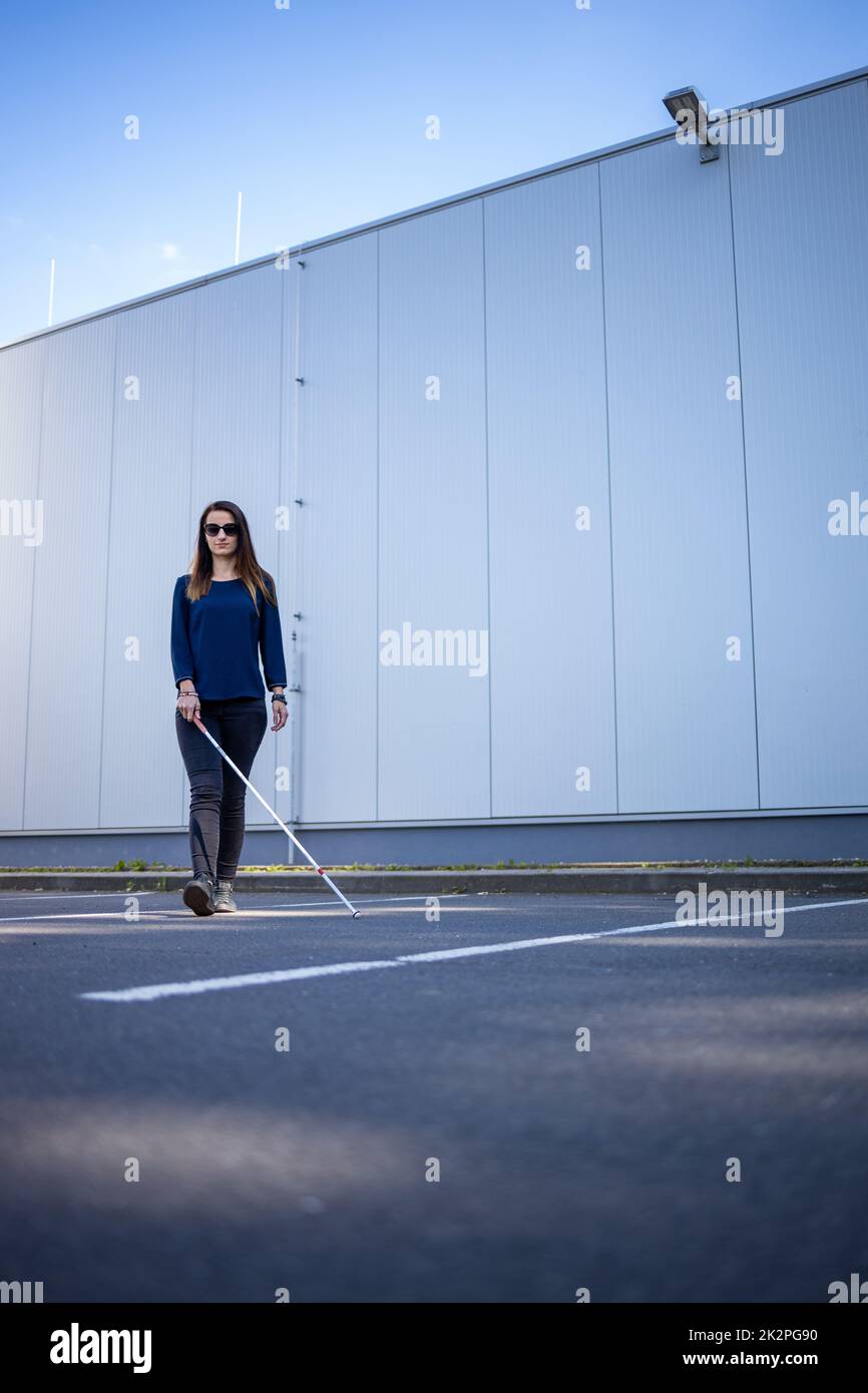 Young woman with impaired vision walking on city streets, using her white cane to navigate the urban space better and to get to her destination safely Stock Photo