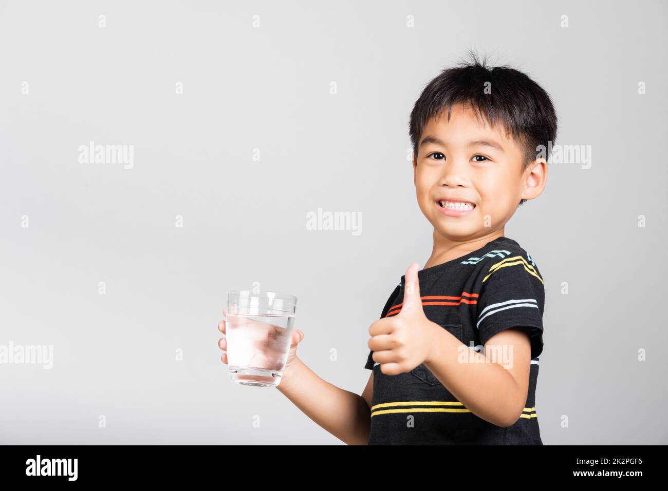 Little cute kid boy 5-6 years old smile drinking fresh water from glass and show thumb up finger for good sign Stock Photo
