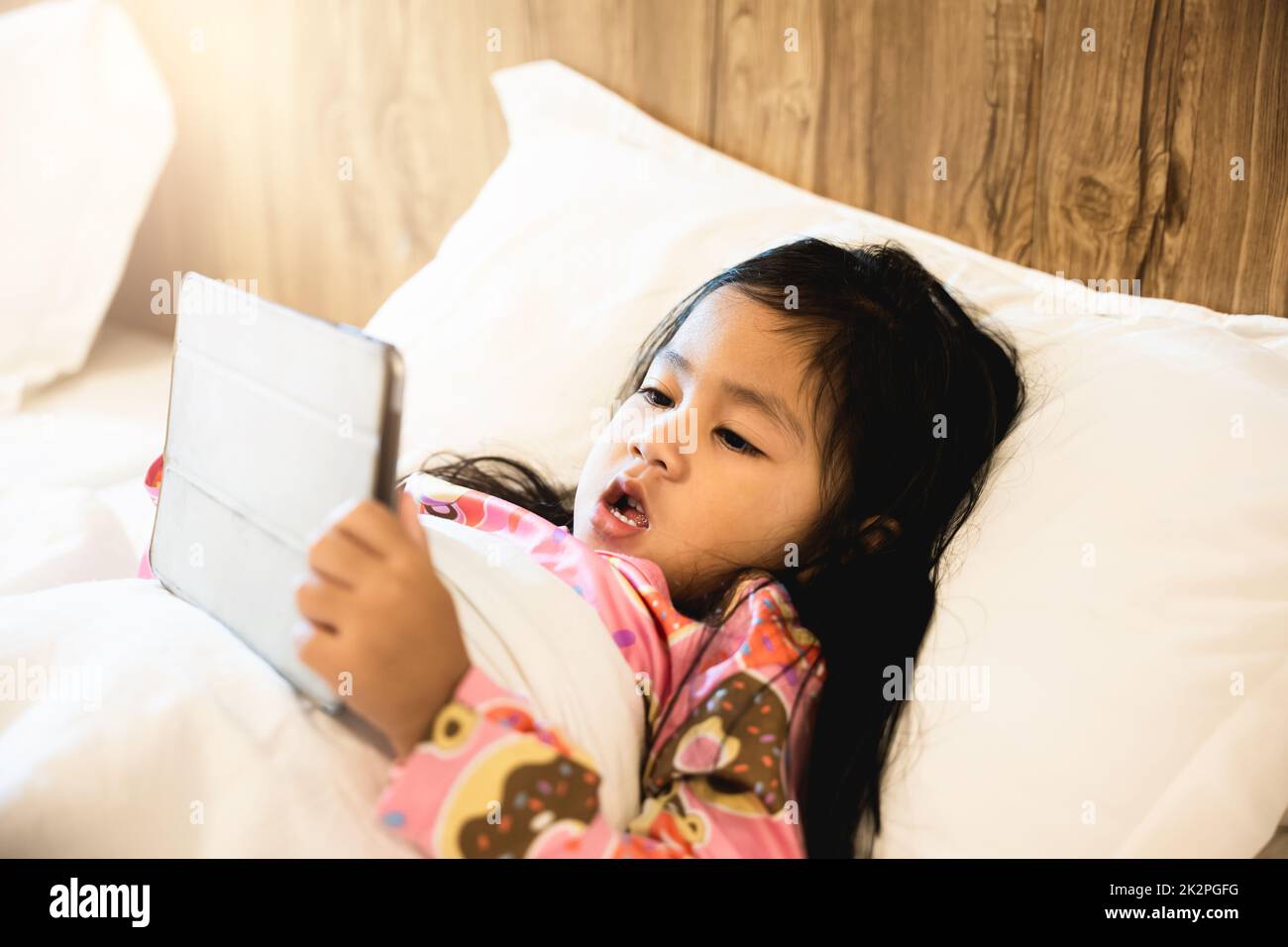 Little Asian girl playing digital tablet on bed addicted game and cartoon Stock Photo