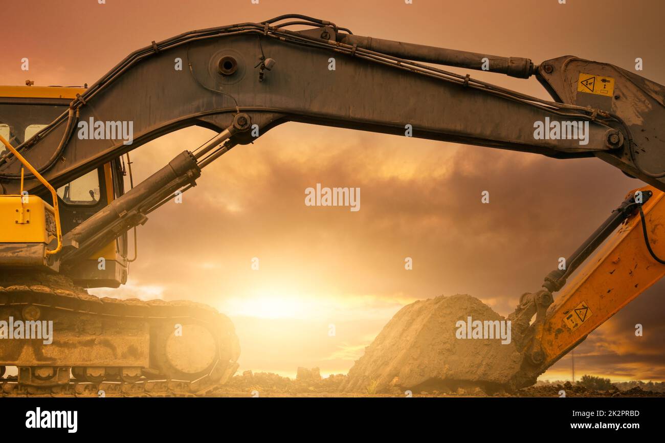 Backhoe parked at construction site after digging soil. Bulldozer on sunset sky and clouds background. Digger after work. Earth moving machine at construction site at dusk. Digger with dirt bucket. Stock Photo
