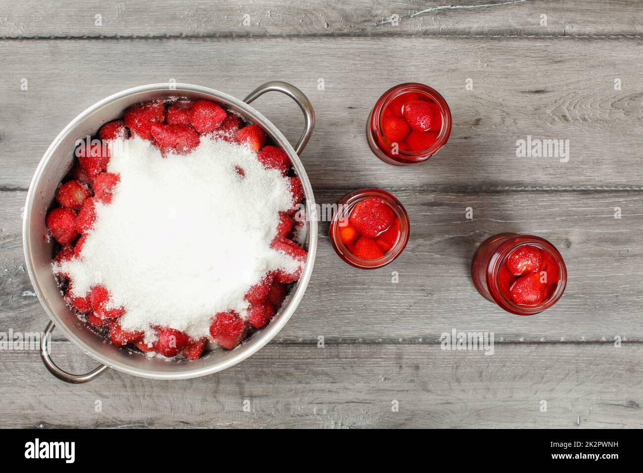Tabletop view, large steel pot full of strawberries covered with crystal sugar, three bottles of strawberry in syrup next to it on gray wood desk. Homemade compote preparation. Stock Photo