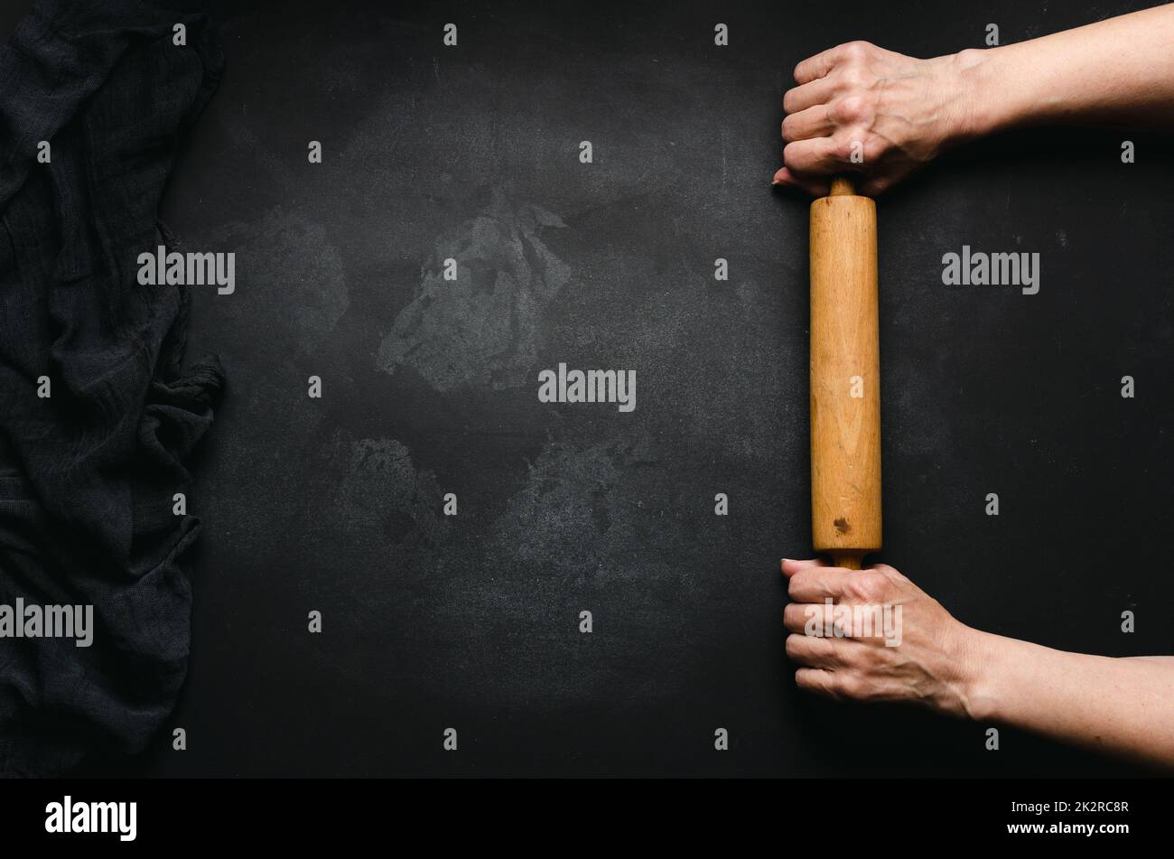 Two female hands hold a wooden rolling pin for rolling dough on a black table, top view. Place for an inscription Stock Photo