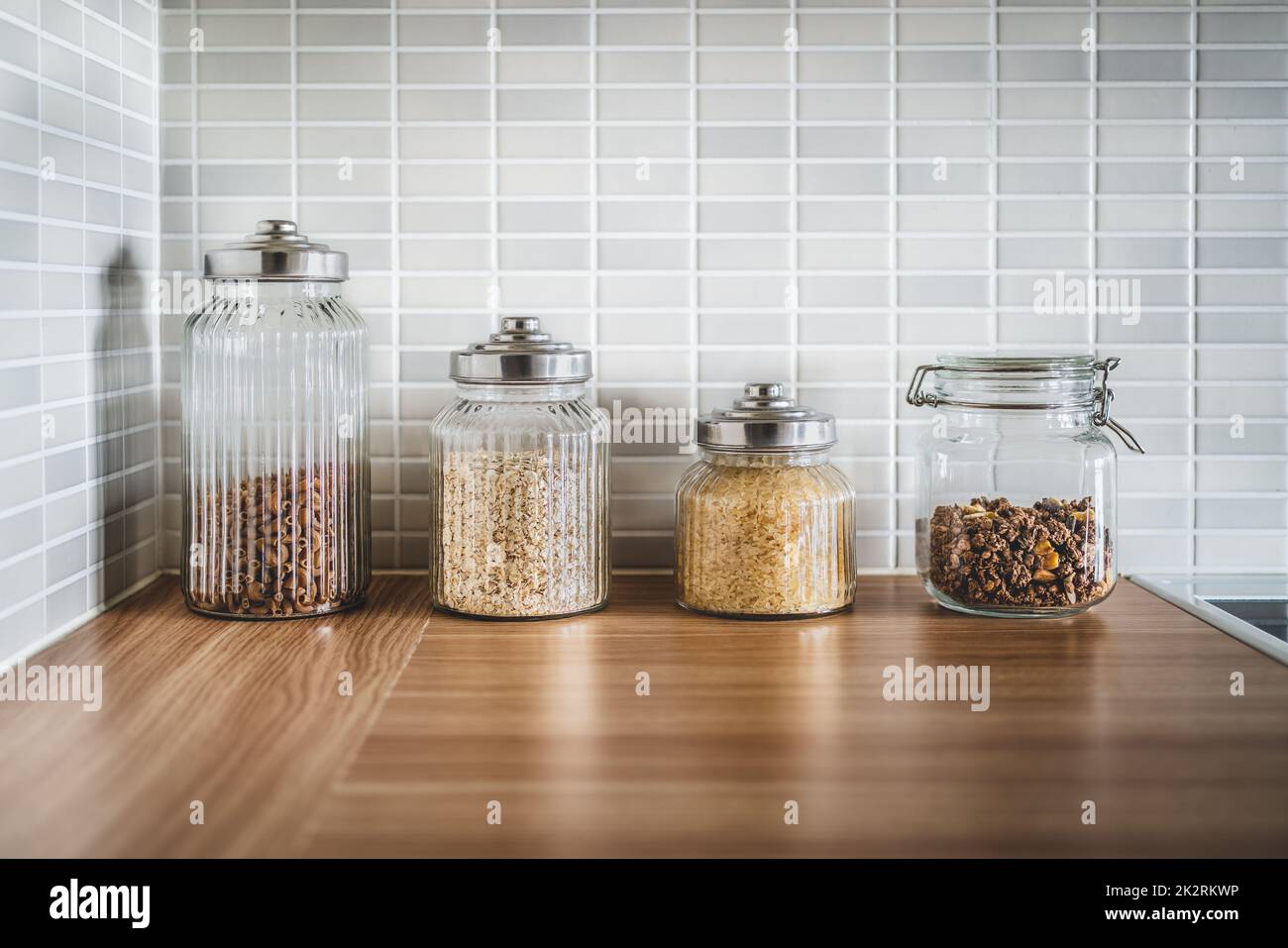 Kitchen containers and food in glass jars. Ingredient organization and sustainable storage. Pasta, oatmeal, rice and muesli. Close up of wood table. Stock Photo