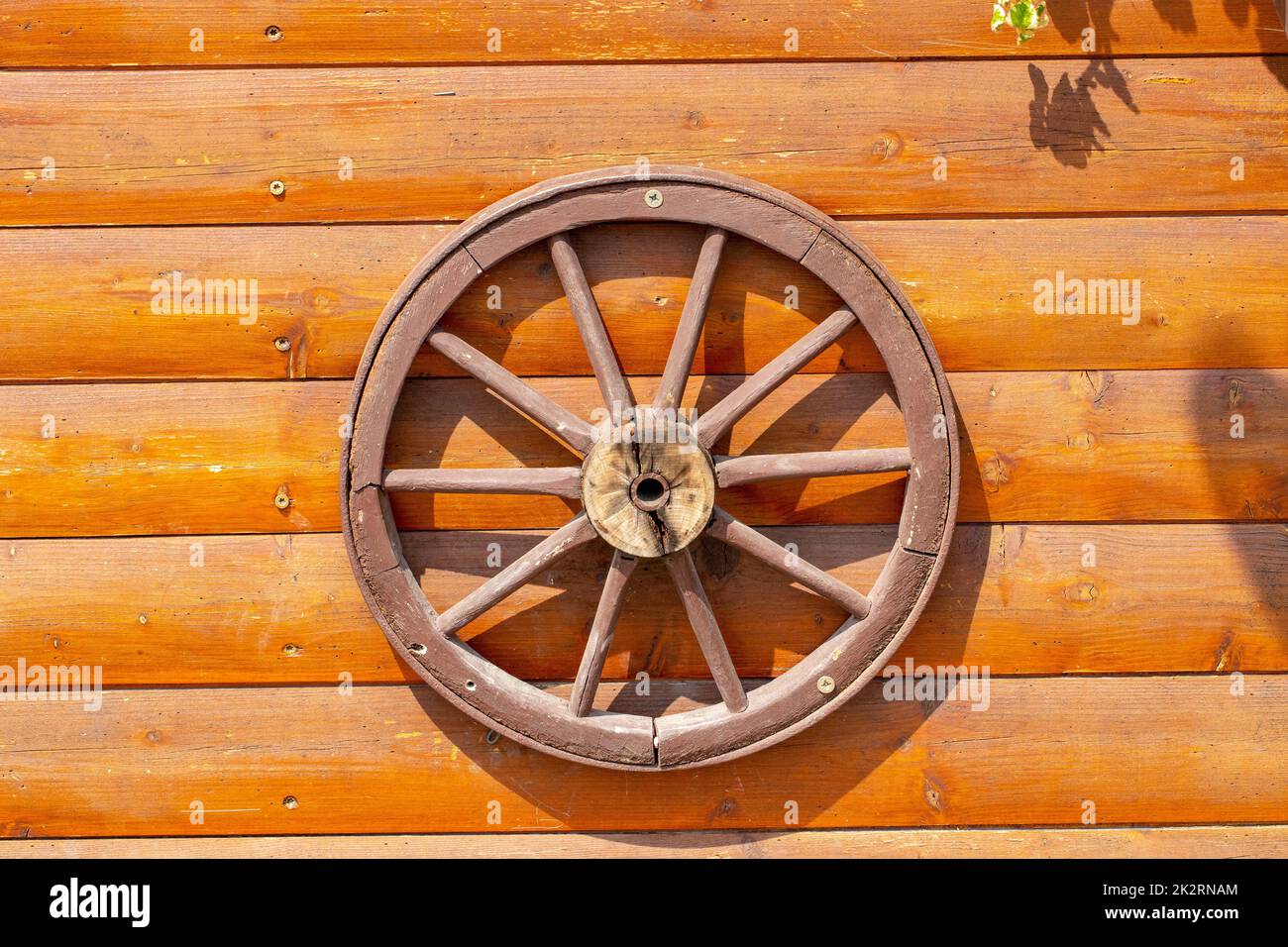 An old wooden carriage wheel with metal fittings hangs on the wall of a wooden house for decoration. Wooden spoke wheel. Outdoor natural light. Macro. Stock Photo