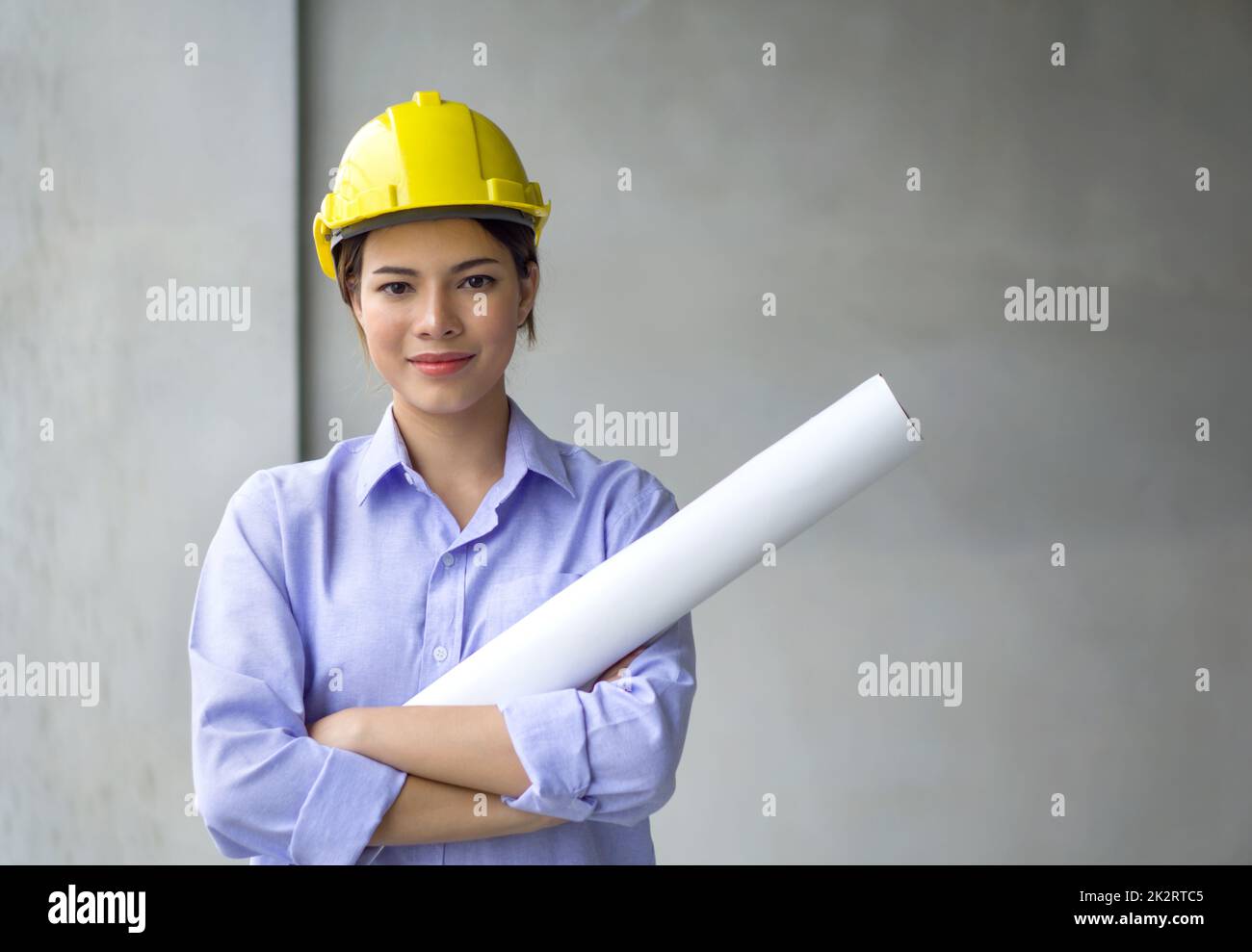 Young woman in construction helmet and floor plan stand with arms crossed in front of gray plaster wall. Work environment of engineers at the construction site of housing projects. Stock Photo