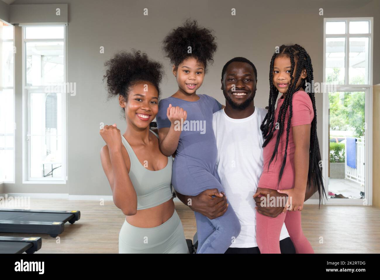 Happy family enjoy holiday together in fitness center. Young father hold both of his daughter in arms while the mother raise her fist up. Stock Photo