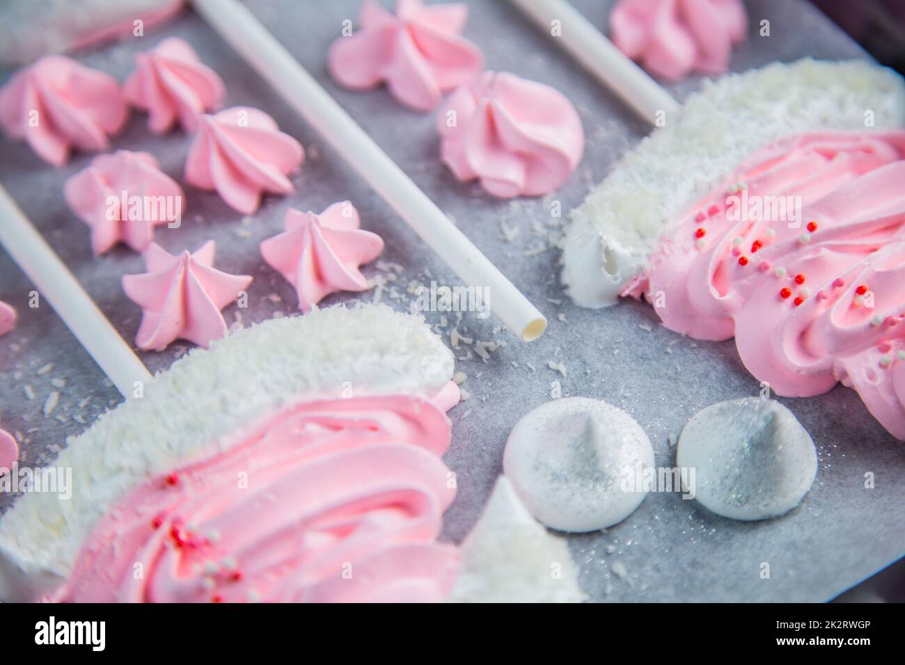 Meringues in the form of pink Christmas hats with a pompom and a white lapel on a stick lie on white parchment in two rows, pink stars and white meringues lie between them Stock Photo