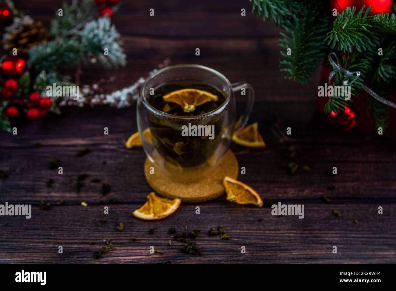 A transparent cup of tea stands on a dark table, on a wooden stand, dried orange slices float in the cup, citrus slices lie next to it, tea is scattered, a photo in a dark key Stock Photo