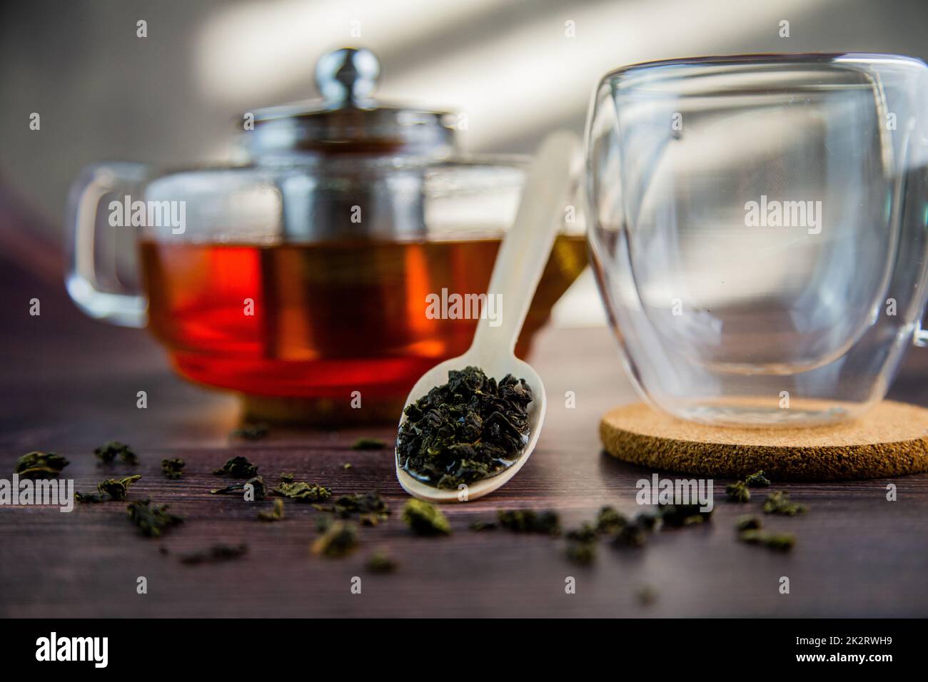 A wooden spoon with tea lies against a dark wood background, a mug and a teapot are visible from behind, a photo in dark colors. Stock Photo