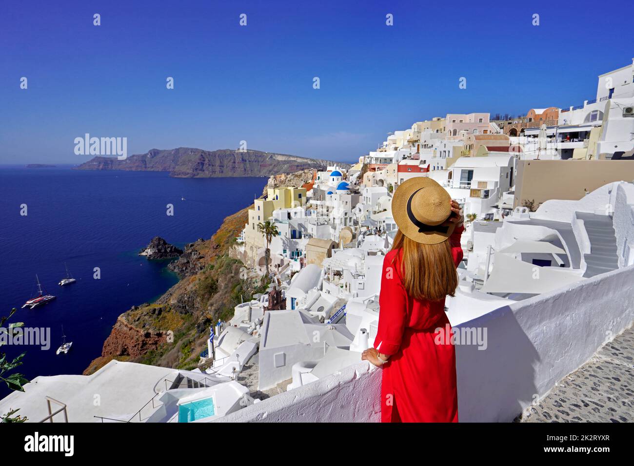 Beautiful woman in Oia village holds hat when looking cityscape from terrace in Santorini Island, Greece Stock Photo