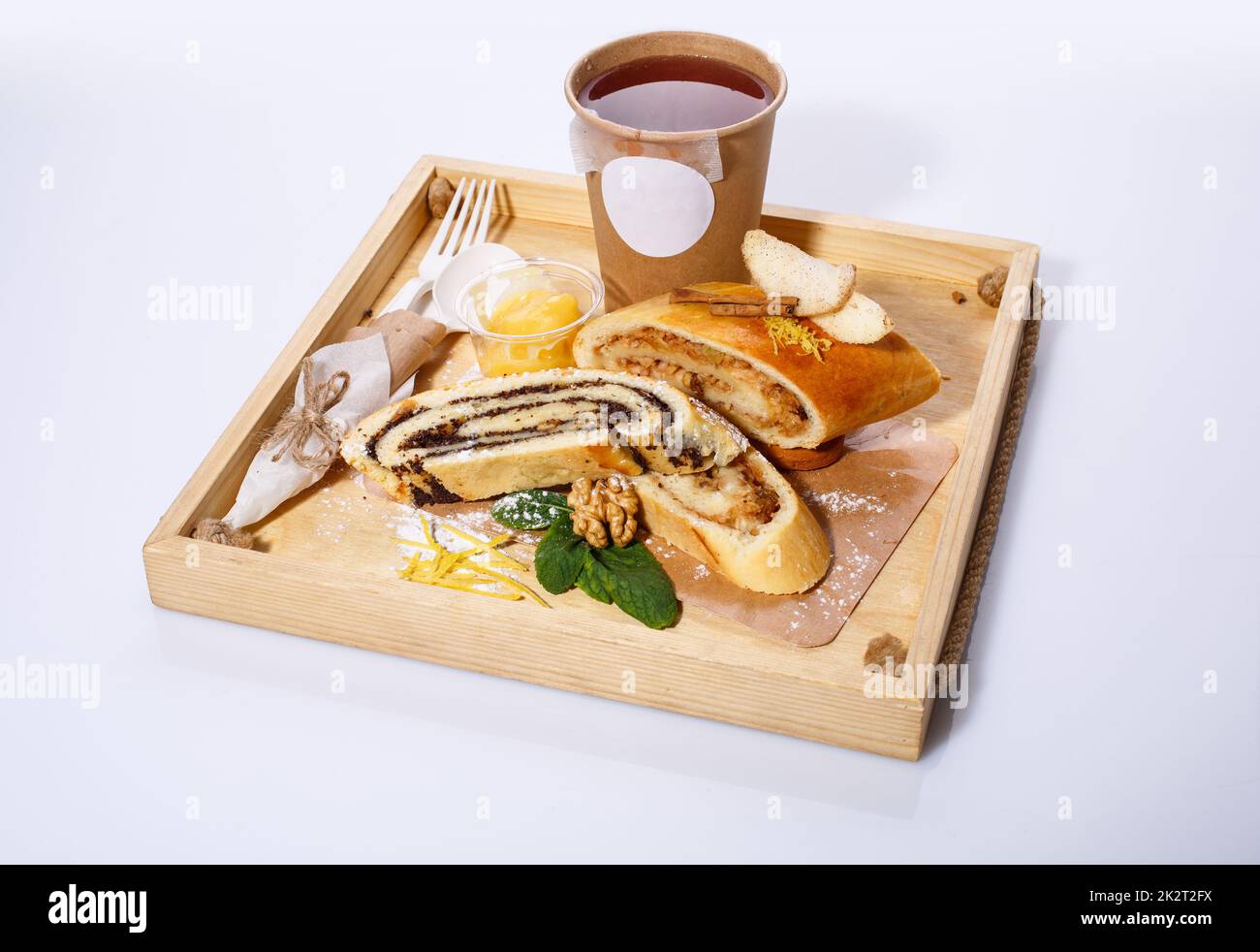 Tea in paper cup, disposable spoon and fork, poppy and apple strudel on a rustic wooden tray. Stock Photo