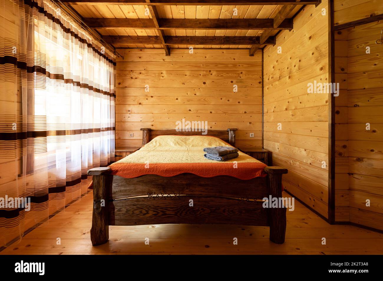 Rustic wooden bed in a cozy cottage room with a double bed, bedside table, blankets and towels. Stock Photo