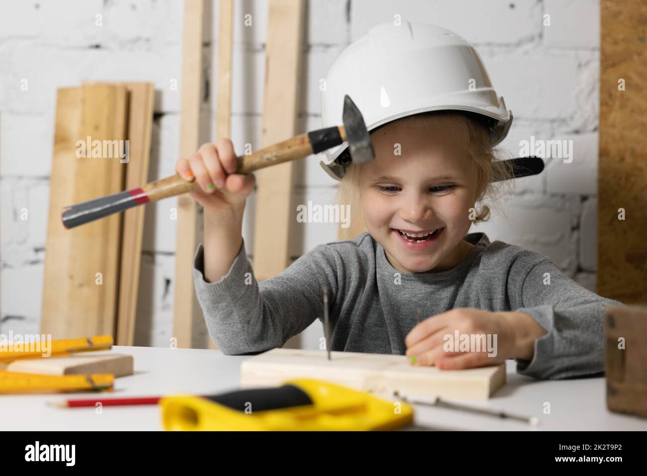 happy little girl with helmet try to hammer a nail in workshop. craft education Stock Photo