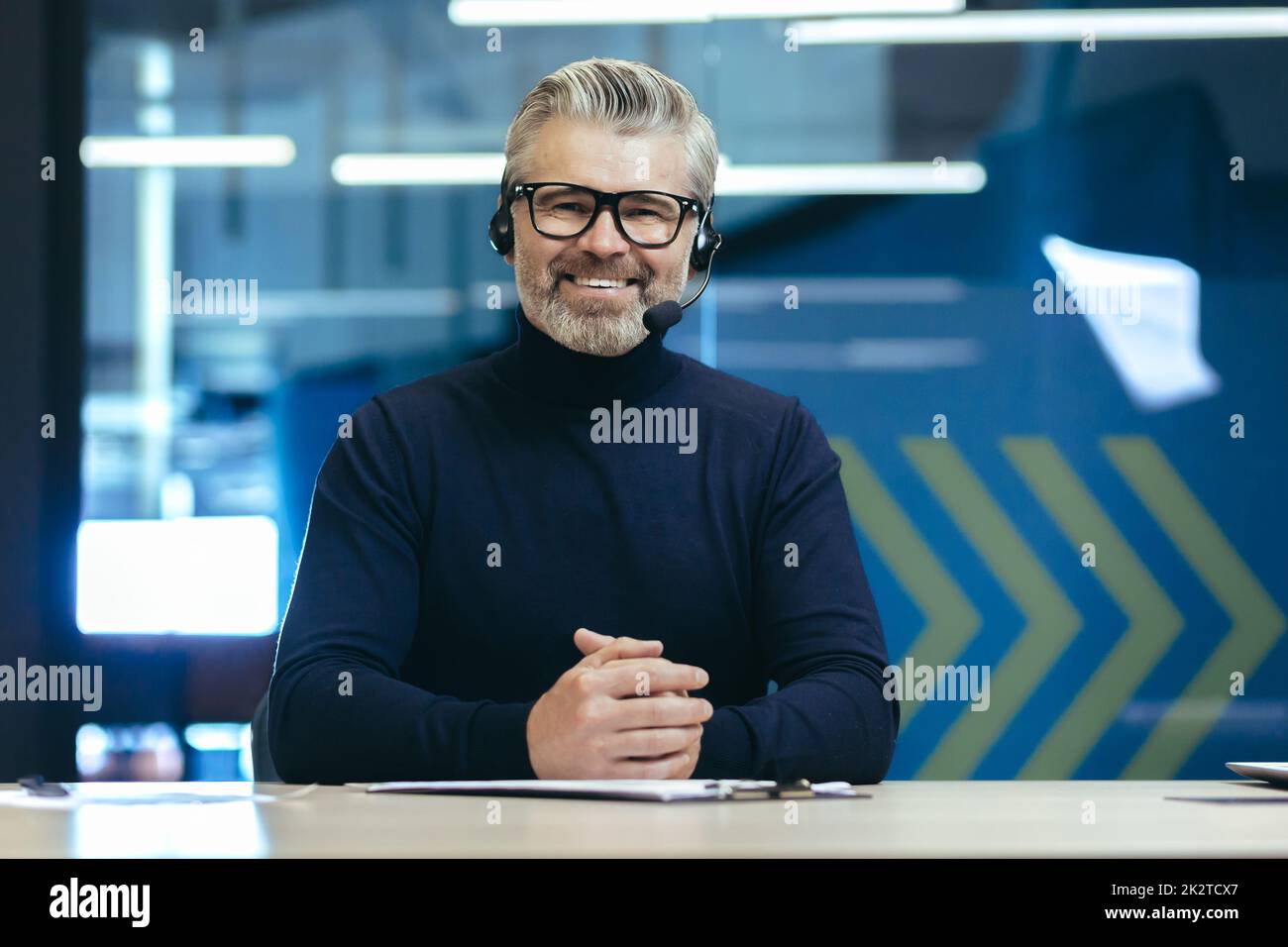 Webcam view, senior experienced gray haired businessman looking at camera and smiling, online meeting, video call man with headset working inside modern office building. Stock Photo