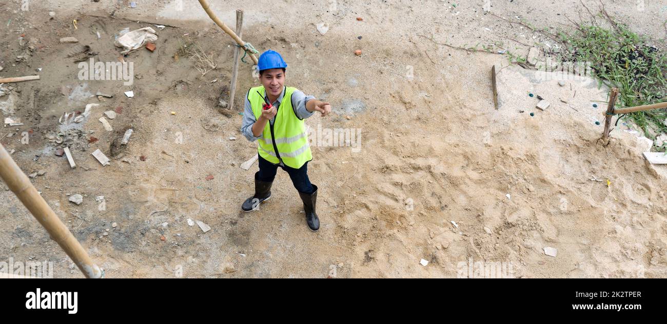 à¹à¹Young asian man in hardhat and  safety vest pointing at scaffolding made of wood structure while holding Walkie-Talkie. Day time work safety checks. Work environment at the site of housing projects. Stock Photo