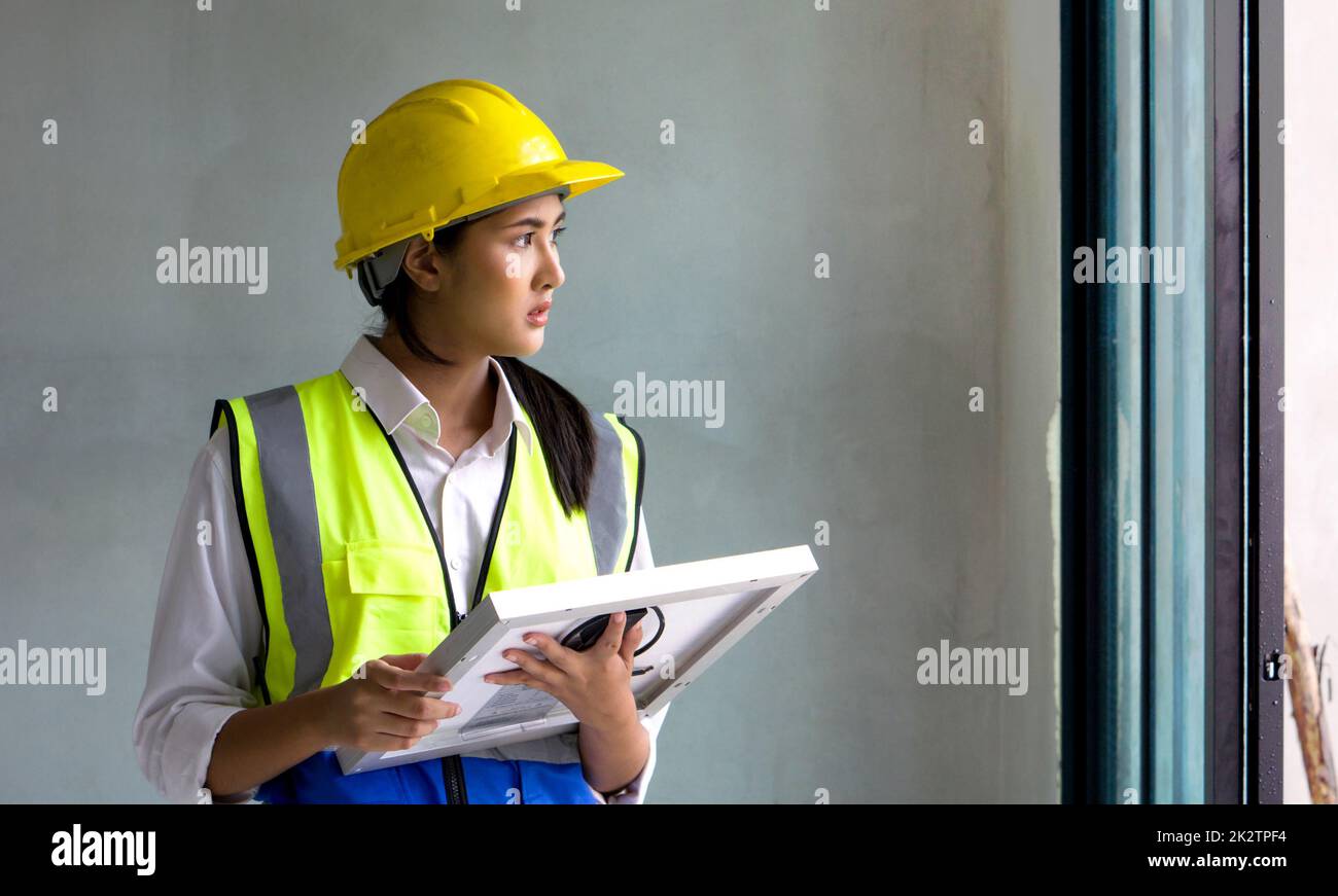 Young asian woman in a safety vest and hardhat holding solar cell panel while looking out of the window. Work environment of engineers at the construction site. Stock Photo