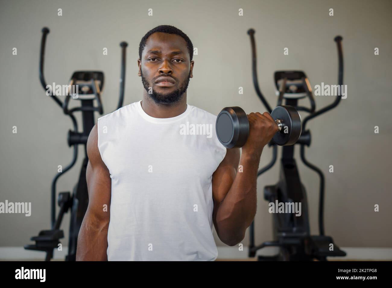 Young short curly black hair man with moustache and beard working on his biceps, lifting barbell with one hand. There are cardio machines in the gym. Stock Photo