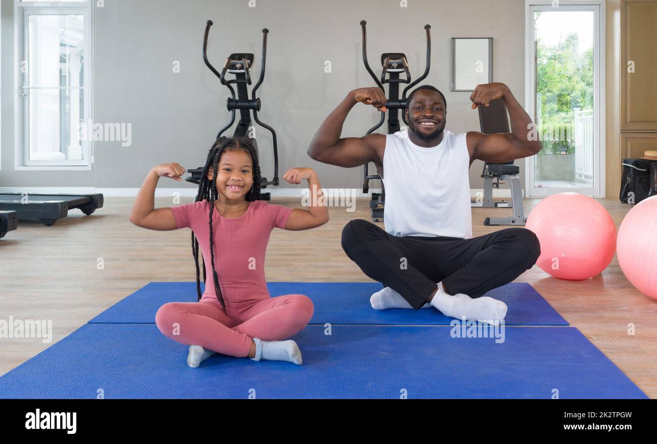 à¸ºBlack Cornrow Braids girl in sportswear flexing muscles, biceps, triceps with her father on yoga mat. Two yoga ball are placed next to them. Happy family enjoy holiday together in f Stock Photo